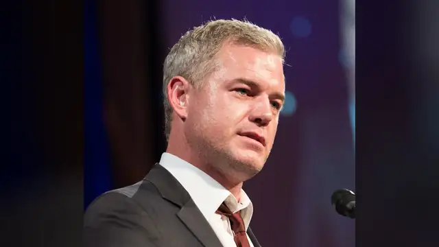 Eric Dane in a dark suit and burgundy tie speaking at a microphone on stage