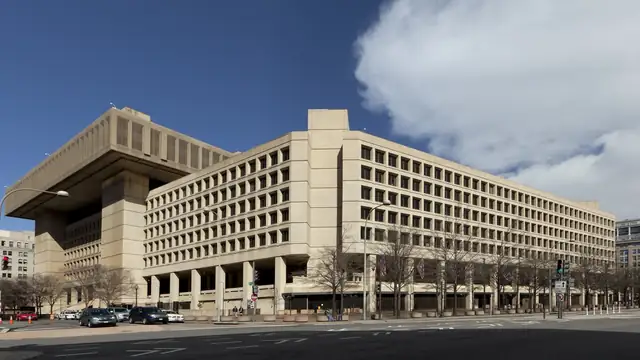 The tan concrete FBI headquarters building on a Washington street corner under a blue sky with a large white cloud