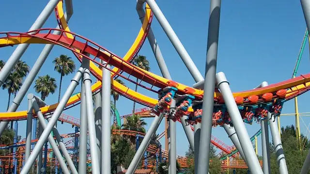 Riders hanging below a red and yellow looping roller coaster among gray steel supports and palm trees