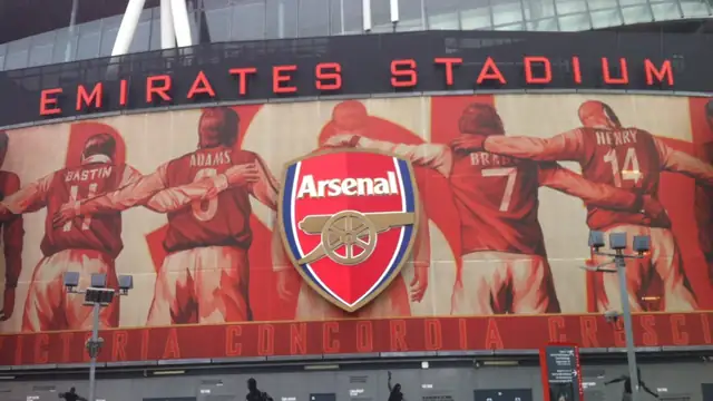 Exterior of the Emirates Stadium in London with the red Arsenal crest and a mural of former players above the entrance