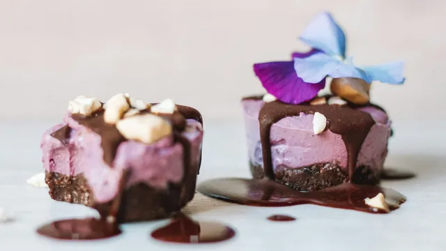 Two mini purple ube cheesecakes with chocolate drizzle, chopped nuts and an edible flower on a light marble surface