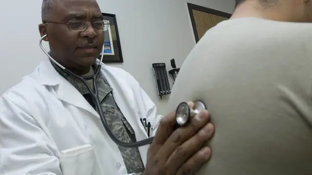 A doctor in a white coat holds a stethoscope to the back of a patient during an exam in a clinic