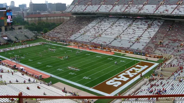 Players warm up on the field at Texas Memorial Stadium in Austin with the burnt orange TEXAS end zone below tall stands and the city skyline