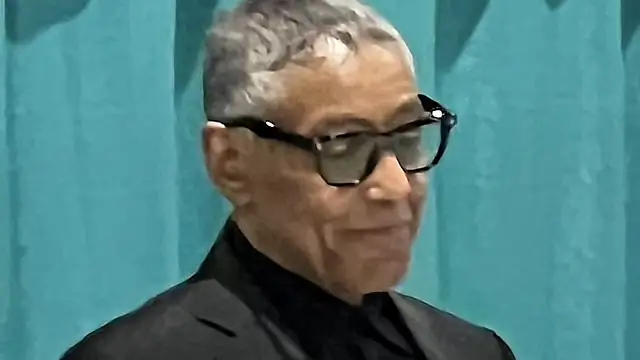 Giancarlo Esposito in dark glasses and a black suit smiling at a fan convention