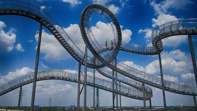 A looping steel track sculpture shaped like a roller coaster under a cloudy blue sky