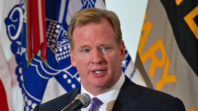 NFL commissioner Roger Goodell in a dark suit and striped tie speaking at a podium in front of flags