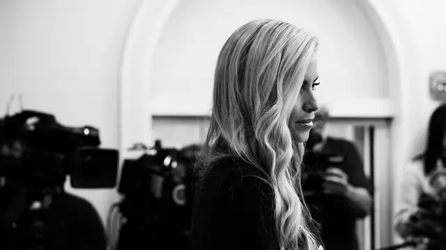 Katie Zacharia in a black and white photo standing at the briefing room podium as TV cameras face her