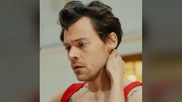 Harry Styles with tousled brown hair touching his neck in a red top