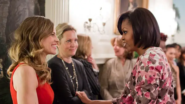 Hilary Swank in a red dress smiling as she greets Michelle Obama at the White House