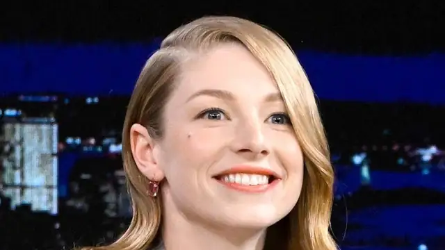 Hunter Schafer smiling with side-swept blond hair against a city skyline backdrop