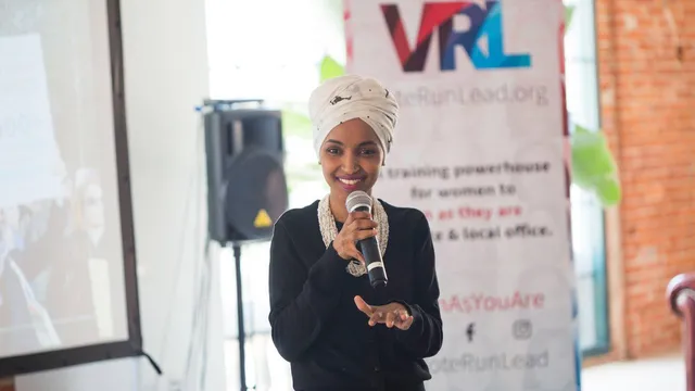Rep. Ilhan Omar in a white head wrap smiling as she speaks into a microphone at a women's leadership training
