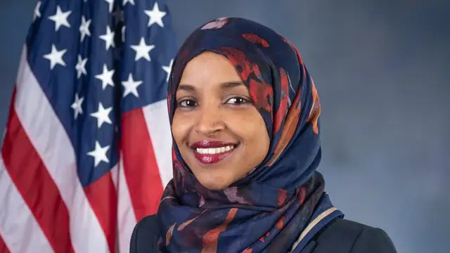 Rep. Ilhan Omar in a navy patterned head scarf smiling in front of an American flag in her official House portrait