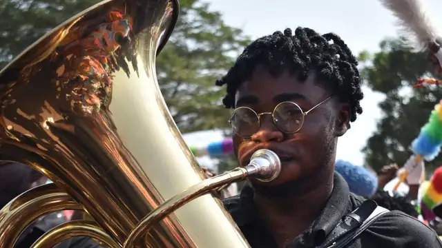 A young Black musician in round glasses playing a shiny brass tuba at an outdoor parade with colorful pom-poms in the background