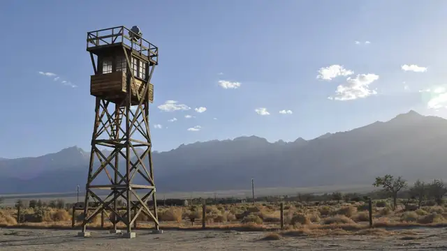A wooden guard tower at the Manzanar incarceration site with mountains behind it under a partly cloudy sky