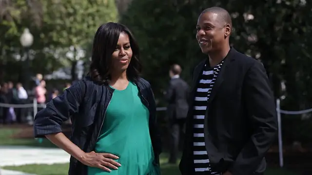 Jay-Z in a black blazer and striped shirt smiles while standing outdoors next to Michelle Obama in a green top on the White House lawn
