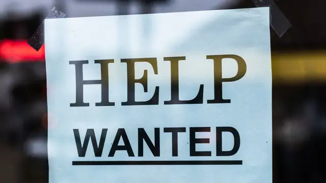 A white help wanted sign hangs in a shop window at night