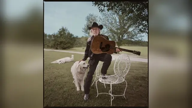 John Anderson in a black cowboy hat holding an acoustic guitar beside a white dog on a green lawn