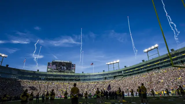 Green Bay Packers players line up on the field before a packed crowd at Lambeau Field as smoke trails from a military flyover streak across a blue sky