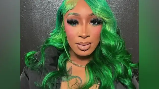 Kash with bright green wavy hair in a black jacket against a dark backdrop