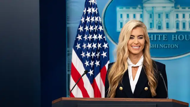Katie Zacharia smiling at the White House briefing room podium beside an American flag