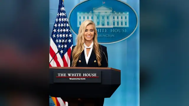 Katie Zacharia smiling in a navy blazer at the White House briefing room podium beside an American flag
