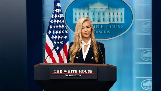 Katie Zacharia with long blonde hair and a dark blazer stands at the White House briefing room podium beside an American flag