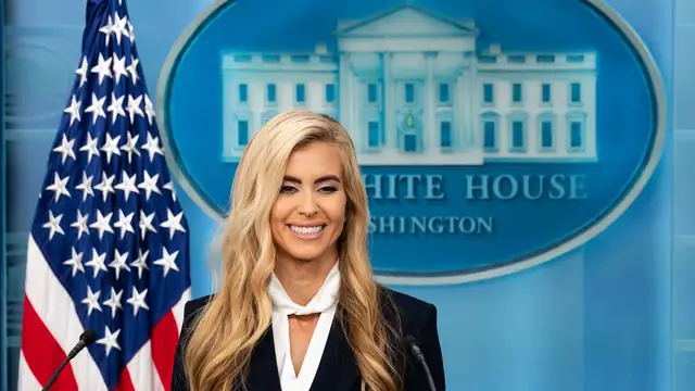 Katie Zacharia smiling in a navy blazer and white bow blouse at the White House briefing room podium