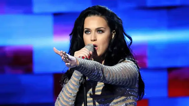 Katy Perry singing into a microphone in a sparkly striped gown on a blue lit stage