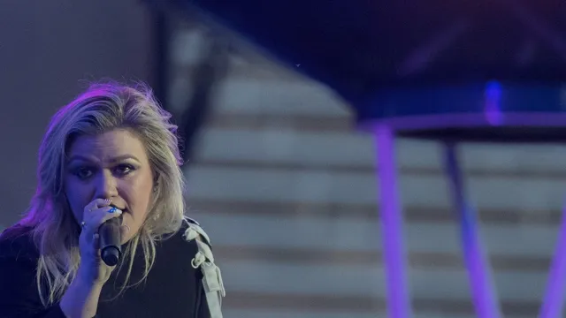 Kelly Clarkson singing into a handheld microphone in a black top with white ribbon trim