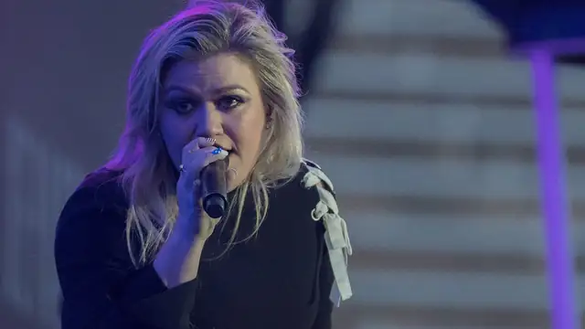 Kelly Clarkson singing into a handheld microphone in a black top with white ribbon trim