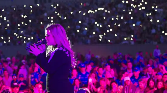 Kelly Clarkson singing into a handheld microphone on stage with a pink-lit crowd below