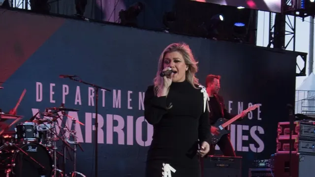 Kelly Clarkson singing into a microphone on an outdoor stage in a black dress