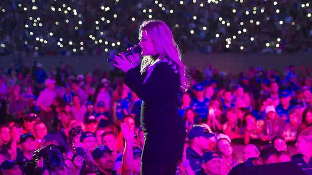 Kelly Clarkson singing into a microphone on a stadium stage in front of a cheering crowd