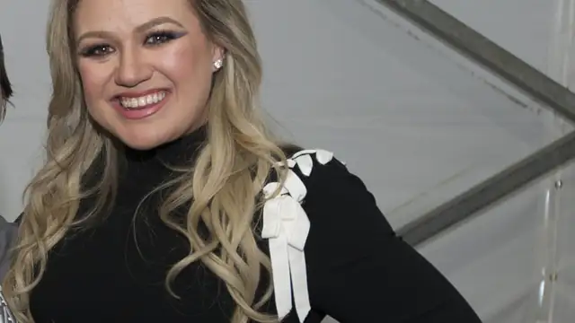Kelly Clarkson smiling in a black top with a white bow at an indoor event