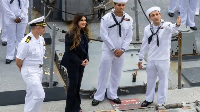 Kimberly Guilfoyle in a dark suit smiling on the deck of a Navy destroyer beside sailors in white dress uniforms
