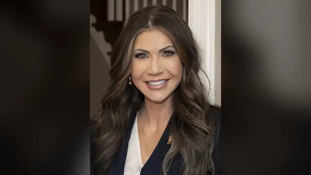 Kristi Noem smiling with long dark wavy hair in a navy blazer and white blouse