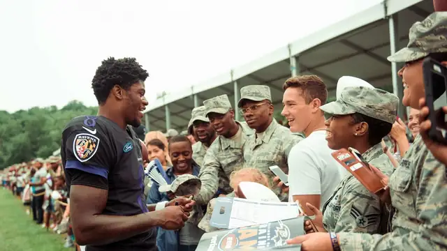 Lamar Jackson in a black Ravens practice jersey smiles as he signs autographs for service members along a rope line at training camp