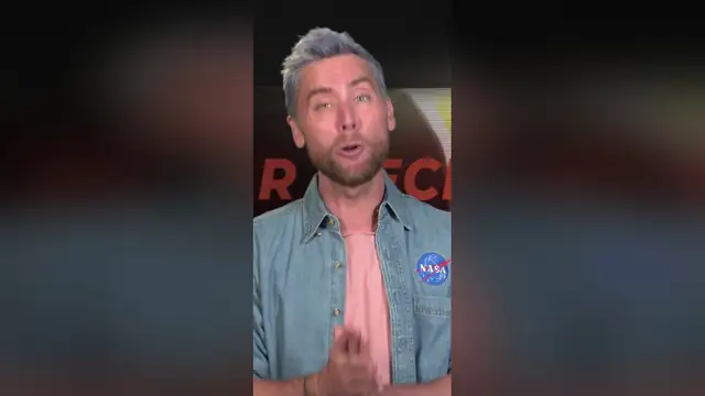 Lance Bass in a denim shirt speaking to the camera in a NASA studio