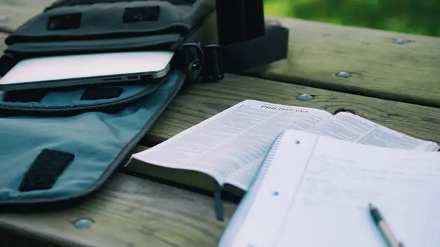 An open notebook and a pen sit on a wooden table beside a laptop bag as a student works on assignments