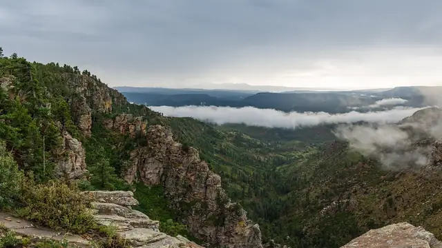Pine-covered cliffs of the Mogollon Rim in Arizona's rim country under a heavy gray storm sky with low clouds drifting through the valley below