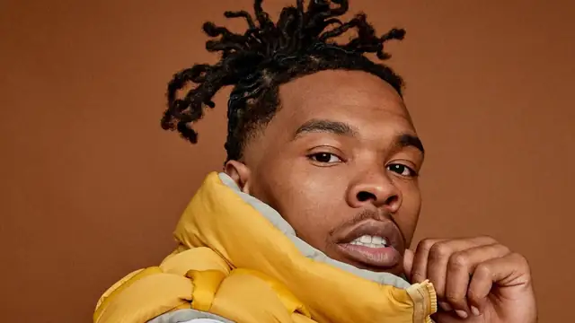 Lil Baby with twisted hair in a yellow puffer vest and one hand at his chin against an orange background