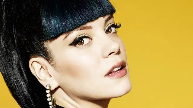 Lily Allen with dark glossy bangs and gold eye makeup looking over her shoulder against a yellow backdrop