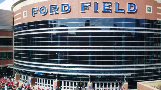The curved glass facade of Ford Field in Detroit under the stadium's blue lettered sign with fans gathered below