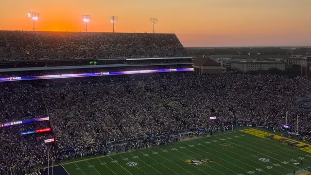 Fans fill LSU's Tiger Stadium under an orange sunset sky with the lit green field and purple end zone below
