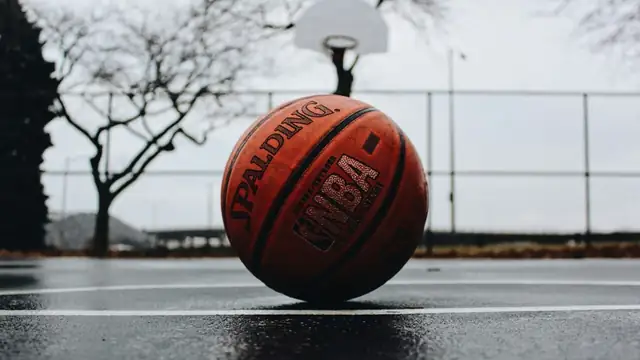 A worn orange Spalding basketball rests on a wet outdoor court with a hoop blurred in the background