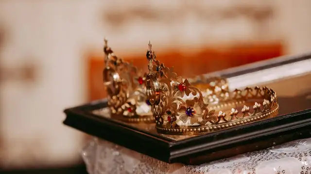 Two ornate gold crowns set with colored stones resting on a dark tray over a white lace cloth