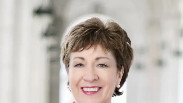 Sen. Susan Collins smiling in a red jacket in her official Senate portrait with a blurred Capitol interior behind her