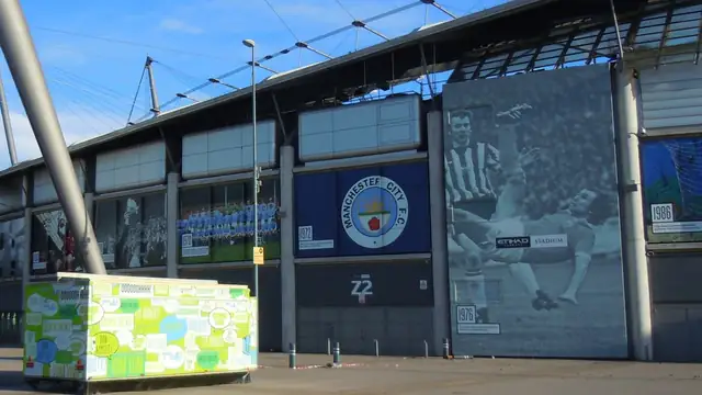 The blue Manchester City club crest on the side of the Etihad Stadium next to a giant black and white mural of a past City player under a bright blue sky