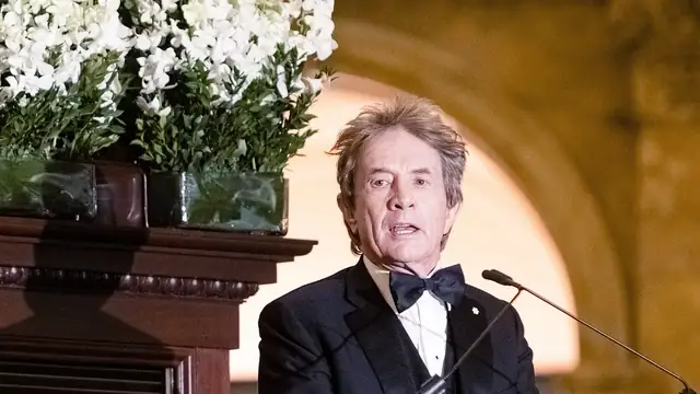 Martin Short in a tuxedo and bow tie speaking at a podium beside white flowers