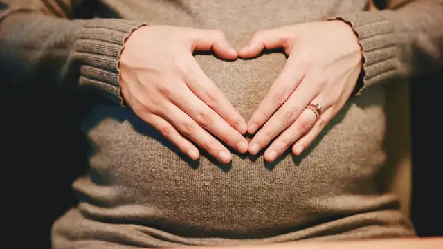 A pregnant woman in a brown sweater cradling her belly with both hands shaped into a heart